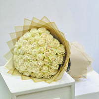 A large bouquet of cream-colored roses wrapped in sheer beige tissue paper, displayed on a white pedestal against a light gray background.