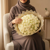 A large bouquet of cream-colored roses wrapped in sheer beige tissue paper, displayed on a white pedestal against a light gray background.