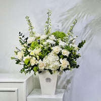 A large artificial flower arrangement in a white rectangular vase, featuring white roses, hydrangeas, green hydrangea blooms, and green foliage, displayed on a white pedestal against a light gray wall.