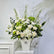 A large artificial flower arrangement in a white rectangular vase, featuring white roses, hydrangeas, green hydrangea blooms, and green foliage, displayed on a white pedestal against a light gray wall.