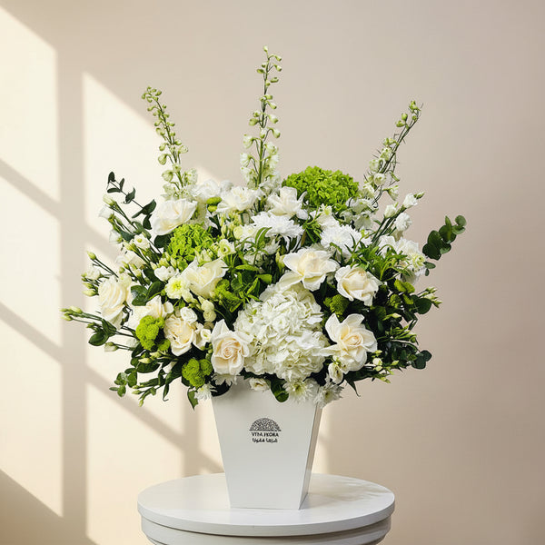 A large artificial flower arrangement in a white rectangular vase, featuring white roses, hydrangeas, green hydrangea blooms, and green foliage, displayed on a white pedestal against a light gray wall.