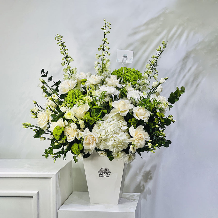 A large artificial flower arrangement in a white rectangular vase, featuring white roses, hydrangeas, green hydrangea blooms, and green foliage, displayed on a white pedestal against a light gray wall.