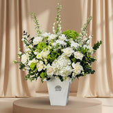 A large artificial flower arrangement in a white rectangular vase, featuring white roses, hydrangeas, green hydrangea blooms, and green foliage, displayed on a white pedestal against a light gray wall.