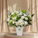 A large artificial flower arrangement in a white rectangular vase, featuring white roses, hydrangeas, green hydrangea blooms, and green foliage, displayed on a white pedestal against a light gray wall.