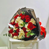 A bouquet of red and cream roses wrapped in black and brown tissue paper with a red ribbon, displayed on a white pedestal against a plain light background.