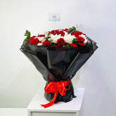 A large bouquet of red and white roses wrapped in black paper with a bright red ribbon, displayed on a white pedestal against a plain light gray wall.