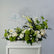 A floral arrangement featuring white calla lilies, white daisies, white roses, and vibrant green pom-pom and globe flowers, displayed in a white rectangular planter box against a light gray wall.