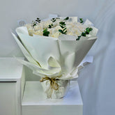 A bouquet of white roses wrapped in white paper with green foliage accents, tied with a gold string, displayed on a white pedestal against a light gray background.