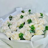 A bouquet of white roses wrapped in white paper, accented with small green eucalyptus sprigs, photographed against a clean white background.