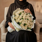 A bouquet of white roses wrapped in white paper with green foliage accents, tied with a gold string, displayed on a white pedestal against a light gray background.