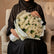 A bouquet of white roses wrapped in white paper with green foliage accents, tied with a gold string, displayed on a white pedestal against a light gray background.
