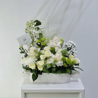 A white floral arrangement featuring white roses, chrysanthemums, green accents, and delicate filler stems in a textured white rectangular vase, displayed on a white pedestal against a light gray background.