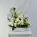 A white floral arrangement featuring white roses, chrysanthemums, green accents, and delicate filler stems in a textured white rectangular vase, displayed on a white pedestal against a light gray background.