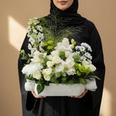 A white floral arrangement featuring white roses, chrysanthemums, green accents, and delicate filler stems in a textured white rectangular vase, displayed on a white pedestal against a light gray background.