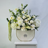 A floral arrangement featuring white and cream flowers with green foliage, presented in a cream-colored ceramic pot with gold trim, set on a white pedestal against a light gray background.