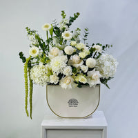 A floral arrangement featuring white and cream flowers with green foliage, presented in a cream-colored ceramic pot with gold trim, set on a white pedestal against a light gray background.