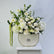 A floral arrangement featuring white and cream flowers with green foliage, presented in a cream-colored ceramic pot with gold trim, set on a white pedestal against a light gray background.