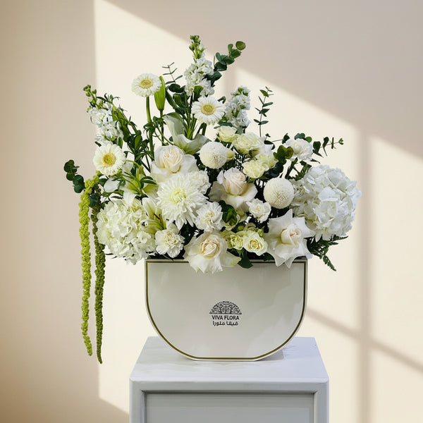 A floral arrangement featuring white and cream flowers with green foliage, presented in a cream-colored ceramic pot with gold trim, set on a white pedestal against a light gray background.