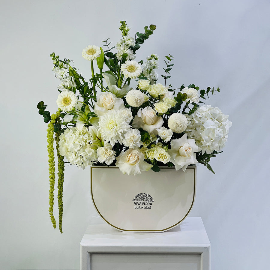 A floral arrangement featuring white and cream flowers with green foliage, presented in a cream-colored ceramic pot with gold trim, set on a white pedestal against a light gray background.