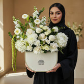 A floral arrangement featuring white and cream flowers with green foliage, presented in a cream-colored ceramic pot with gold trim, set on a white pedestal against a light gray background.