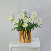 A bouquet of white daisy-like flowers with green stems arranged in a faceted metallic gold vase, placed on a white pedestal against a light gray background.