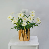A bouquet of white daisy-like flowers with green stems arranged in a faceted metallic gold vase, placed on a white pedestal against a light gray background.