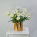 A bouquet of white daisy-like flowers with green stems arranged in a faceted metallic gold vase, placed on a white pedestal against a light gray background.
