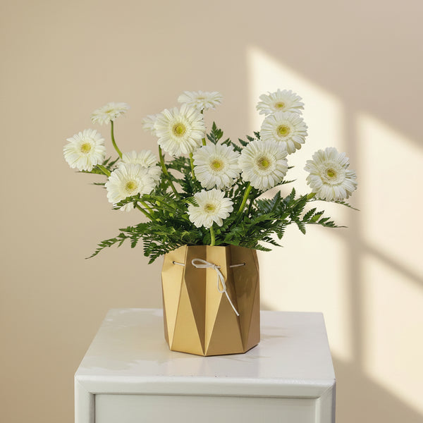 A bouquet of white daisy-like flowers with green stems arranged in a faceted metallic gold vase, placed on a white pedestal against a light gray background.
