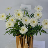 A bouquet of white gerbera daisies with green foliage arranged in a brown paper gift bag, with a small card reading 'VIVA FLOWER DELHI EAST' visible at the top.
