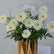 A bouquet of white gerbera daisies with green foliage arranged in a brown paper gift bag, with a small card reading 'VIVA FLOWER DELHI EAST' visible at the top.
