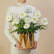A bouquet of white daisy-like flowers with green stems arranged in a faceted metallic gold vase, placed on a white pedestal against a light gray background.
