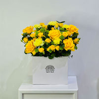 A vibrant yellow flower box arrangement featuring yellow roses, yellow chrysanthemums, and green foliage, presented in a white square box with the brand 'VIVA FLORA' printed on the front, sitting on a white table against a light gray wall.