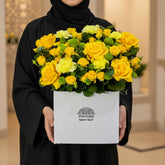 A vibrant yellow flower box arrangement featuring yellow roses, yellow chrysanthemums, and green foliage, presented in a white square box with the brand 'VIVA FLORA' printed on the front, sitting on a white table against a light gray wall.