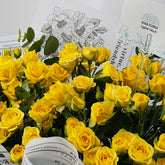 A dense bouquet of bright yellow roses with green leaves, arranged with printed cards and a small gold paperclip in the background.