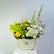 A floral arrangement in a cream-colored ceramic pot with gold trim, featuring yellow roses, white carnations, green hydrangeas, and tall white delphiniums, displayed on a white pedestal against a light gray background.