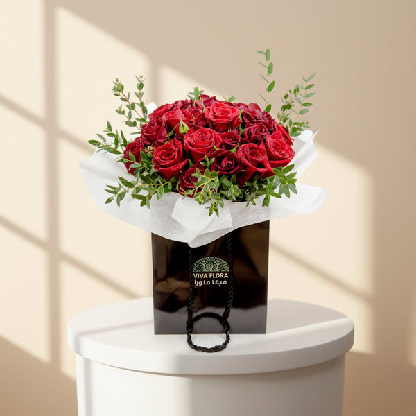 A bouquet of red roses with green leaves, wrapped in white paper and delivered in a black bag with a Viva Flora label.