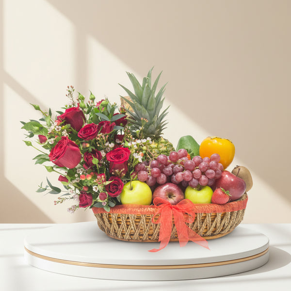 A fruit basket containing apples, kiwi, grapes, strawberries, orange, avocado, kaki, and pineapple, decorated with white and red flowers, presented in a brown wicker basket with a red ribbon.