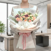 A woman in a beige blazer holds a large, beautifully wrapped flower bouquet in a bright, modern office setting. The bouquet features a mix of peach, pink, white, and burgundy roses with green foliage, wrapped in white paper with a soft pink ribbon.