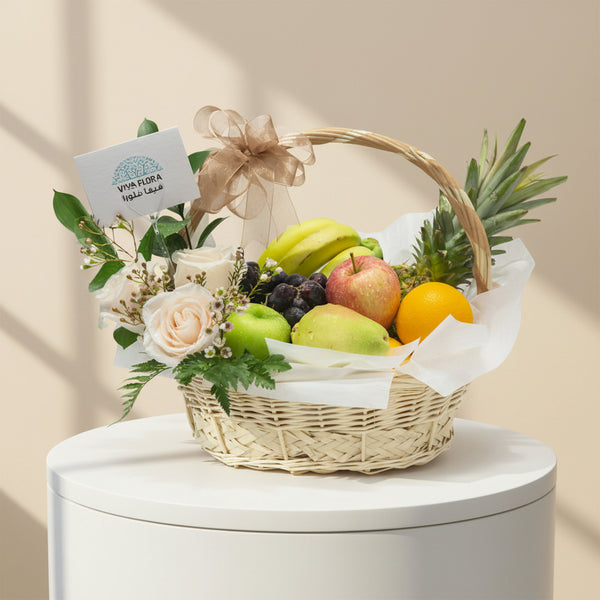 A wicker basket containing a variety of fresh fruits including apples, pears, banana, grapes, orange, and pineapple, decorated with white flowers and greens.