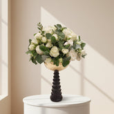 A bouquet of white roses and green foliage arranged in a gold bowl on a dark pedestal stand, displayed on a white round table in a softly lit room.