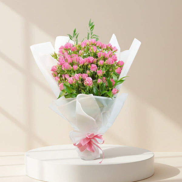 A bouquet of pink flowers wrapped in white tissue paper and tied with a pink ribbon, displayed on a round white pedestal against a warm beige background with soft window shadows.