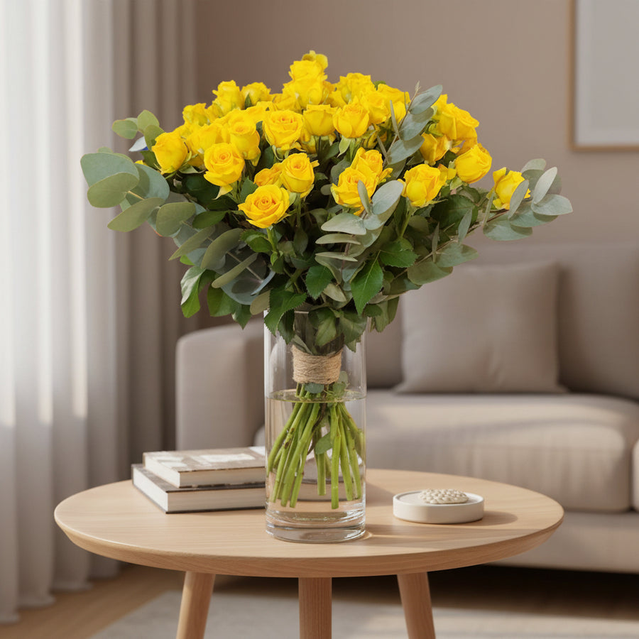 A clear cylinder vase containing a bouquet of yellow roses and green eucalyptus leaves.