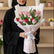 A person wearing a black abaya and hijab holds a wrapped bouquet of pink, red, and white tulips with green leaves and stems, tied with a white ribbon, in a softly lit indoor setting with blurred shelves in the background.