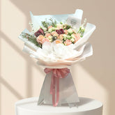 A beautifully arranged floral bouquet with peach, pink, and burgundy roses, wrapped in white tissue paper and a pink ribbon, displayed on a white pedestal against a warm beige background.
