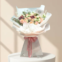 A beautifully arranged floral bouquet with peach, pink, and burgundy roses, wrapped in white tissue paper and a pink ribbon, displayed on a white pedestal against a warm beige background.