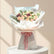 A beautifully arranged floral bouquet with peach, pink, and burgundy roses, wrapped in white tissue paper and a pink ribbon, displayed on a white pedestal against a warm beige background.