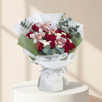 A bouquet of red roses, pink orchids, and green foliage wrapped in white paper and tied with a white ribbon, displayed on a white pedestal against a warm beige background with soft window shadows.
