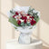 A bouquet of red roses, pink orchids, and green foliage wrapped in white paper and tied with a white ribbon, displayed on a white pedestal against a warm beige background with soft window shadows.