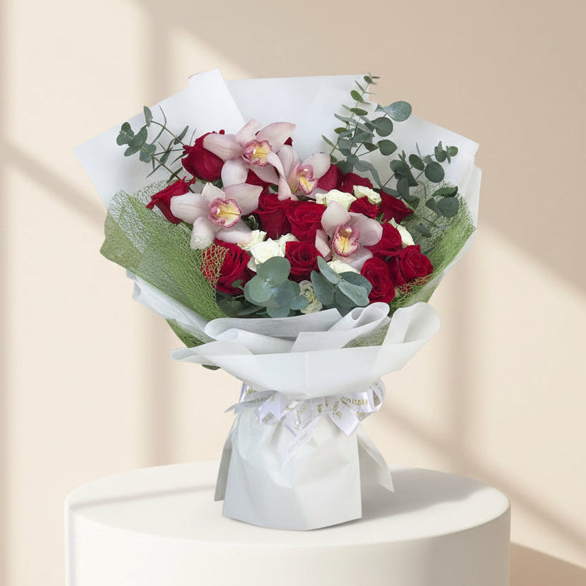 A bouquet of red roses, pink orchids, and green foliage wrapped in white paper and tied with a white ribbon, displayed on a white pedestal against a warm beige background with soft window shadows.